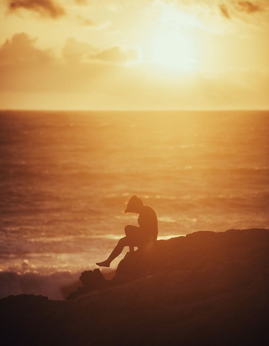 woman with her head down sitting on a rock with the sunset over the ocean